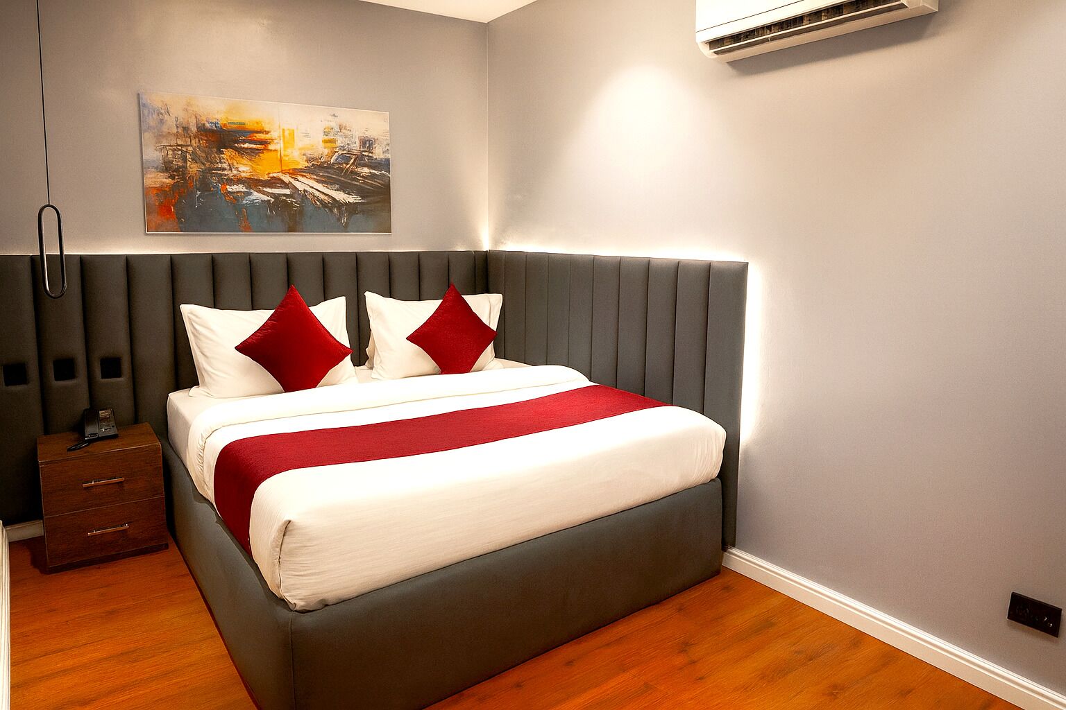 A modern bedroom with a grey upholstered headboard, a white bedspread with red accents, and a painting on the wall.