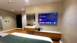 Moderni hotellihuone, jossa on suuri sänky, yöpöytä, jossa on puhelin ja vesipulloja, Google TV -käyttöliittymällä varustettu televisio sekä peili, joka heijastaa huonetta.