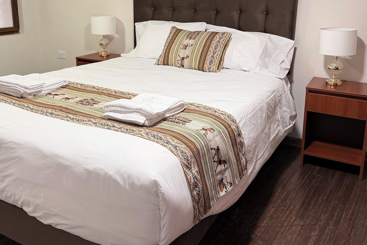 Un lit bien fait, avec une couverture à motifs et des oreillers, une table de chevet avec une lampe et une fenêtre avec des stores.