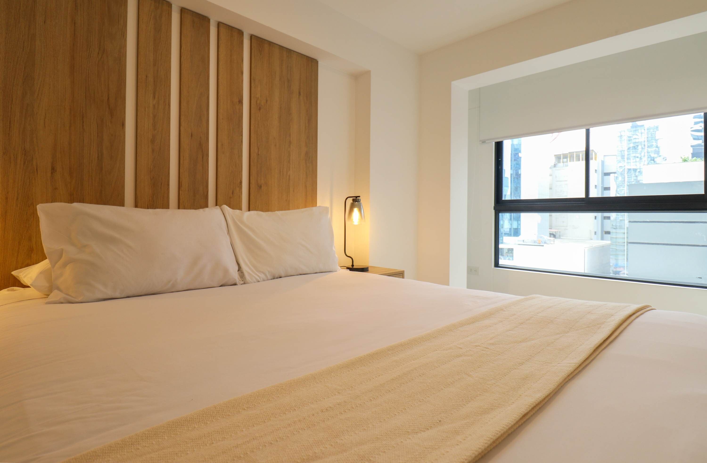 Ett modernt hotellrum med en stor säng, ett träfärgat sänggavel, en sänglampa och ett fönster med utsikt över staden.