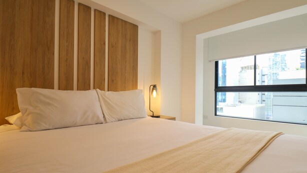 A modern hotel room with a large bed, wooden headboard, bedside lamp, and a window with a city view. - w* | Amazing 1BR Apartment in Heart of San Isidro (San Isidro)