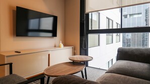 A modern hotel room with a flat-screen TV mounted on the wall, a small round wooden table, a sofa, and a view of the city through large windows. - w* | Amazing 1BR Apartment in Heart of San Isidro (San Isidro)