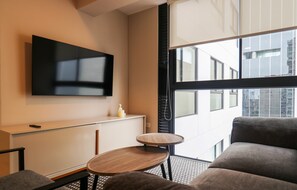 Ein modernes Hotelzimmer mit einem Wand-Fernseher, einem kleinen runden Holztisch, einer Couch und Blick auf die Stadt durch große Fenster.