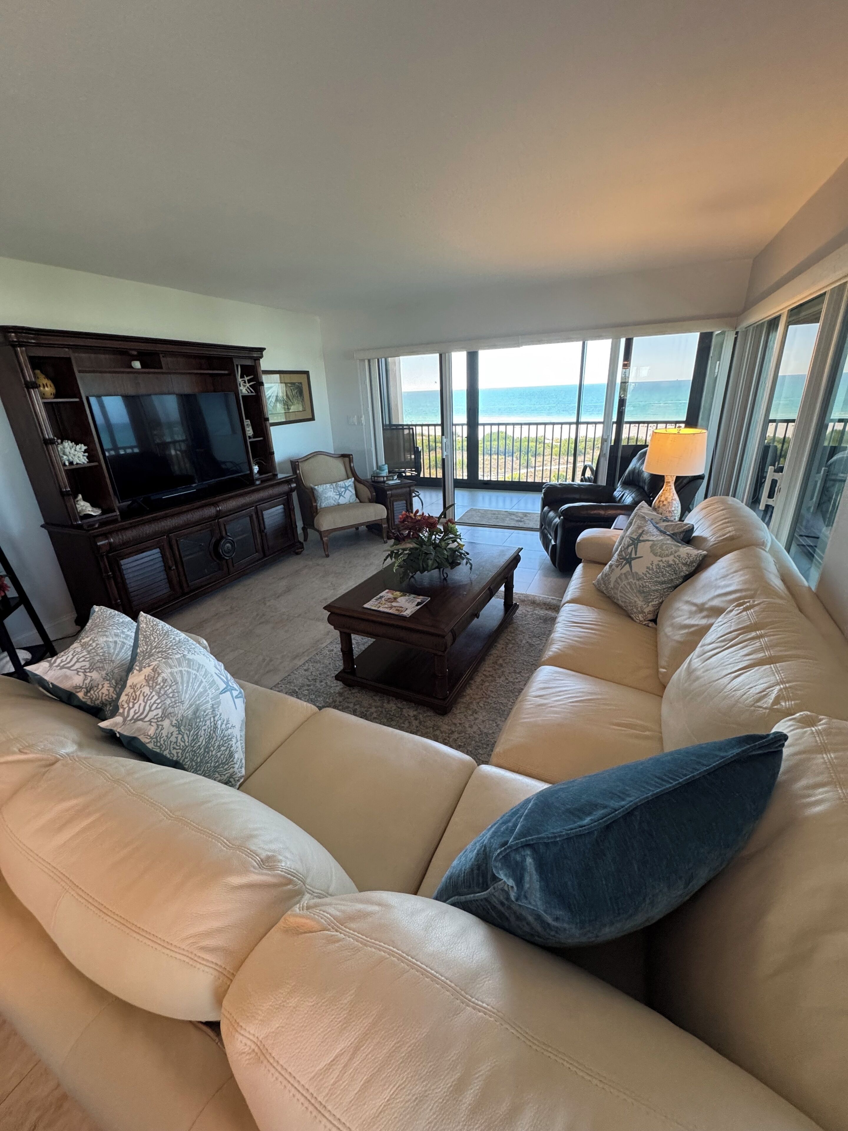 A living room with a large sofa, a coffee table, a TV, and a view of the ocean through large windows.