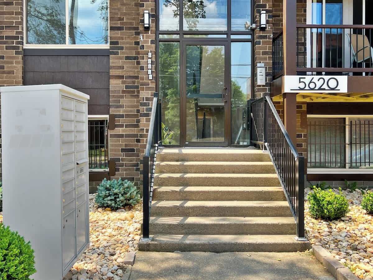 Ein Gebäudeeingang mit Backsteinfassade, einer Glastür und einer Metalltreppe zu einer nummerierten Eingangstür.