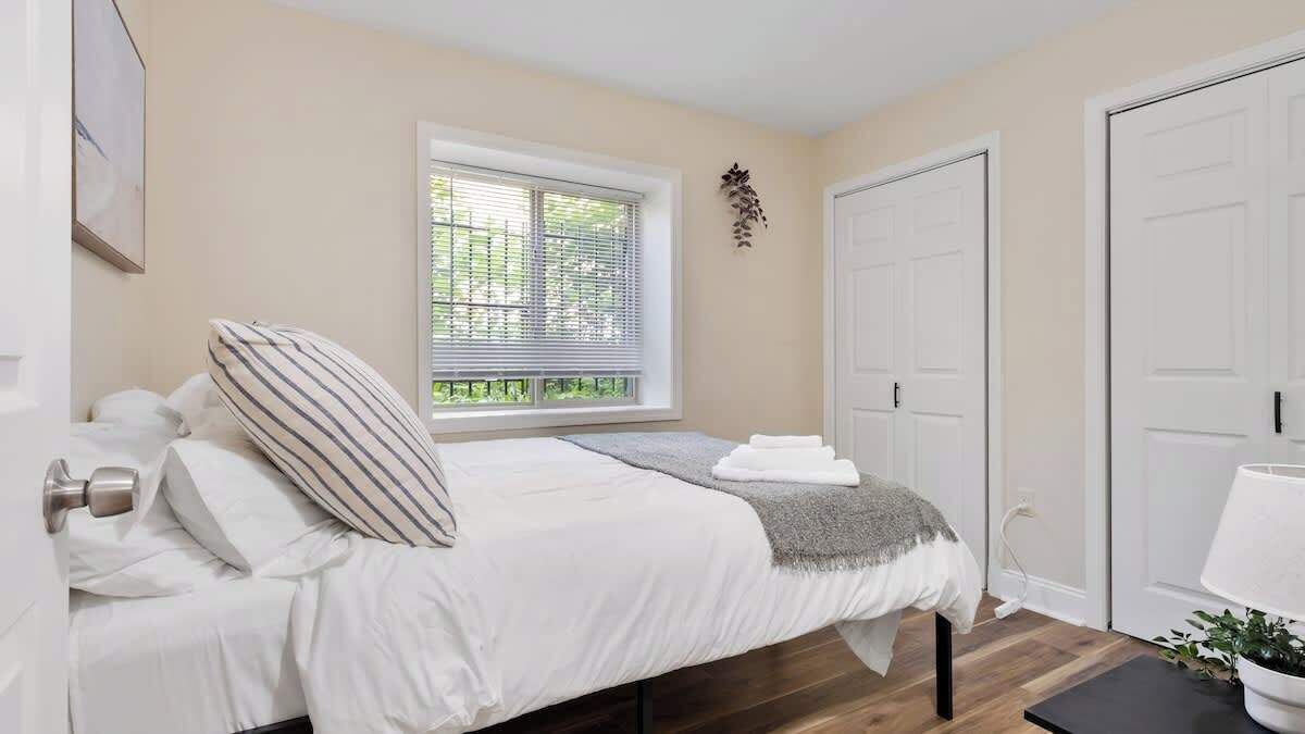 A bedroom with a bed, a window with blinds, a door, and a bedside table with a lamp.