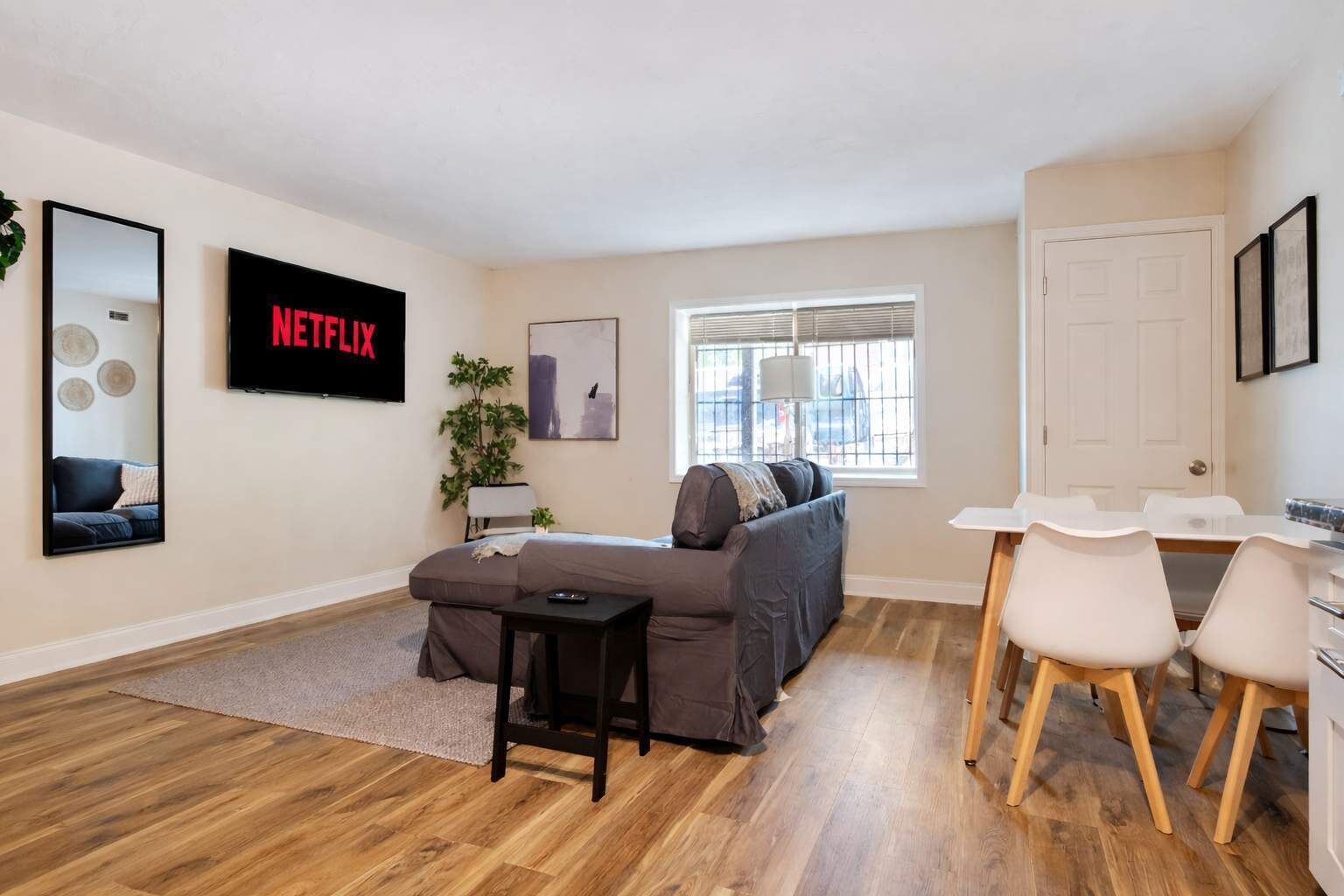 Una sala moderna con un sofá gris, una mesita negra y una mesa de comedor blanca con sillas. Un televisor montado en la pared muestra 'NETFLIX'.