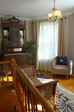 A living room with a wooden staircase, a chandelier, a sofa with a decorative pillow, and a large window with curtains. - The Reflection Suite (Eureka Springs)