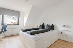 A modern bedroom with a white bed, grey bedding, a blue chair, and a window with a view of a building. - Apartment EDEN - 1 Min HBF - Ideal for trips to Cologne, Düsseldorf, Wuppertal (Solingen)
