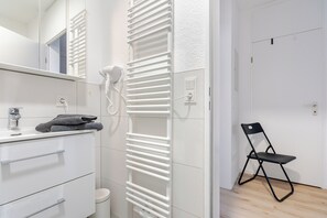 A bathroom with a white vanity, a towel rack, a hairdryer, and a black folding chair. - Apartment EDEN - 1 Min HBF - Ideal for trips to Cologne, Düsseldorf, Wuppertal (Solingen)