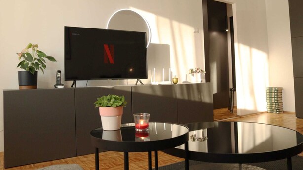 Un salon moderne avec un téléviseur à écran plat posé sur un meuble sombre, une table en verre avec une bougie et une plante en pot.