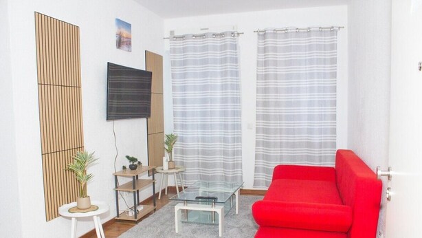 Un salon moderne avec un canapé rouge, une table basse en verre, un téléviseur fixé au mur et une grande fenêtre ornée de rideaux à rayures.
