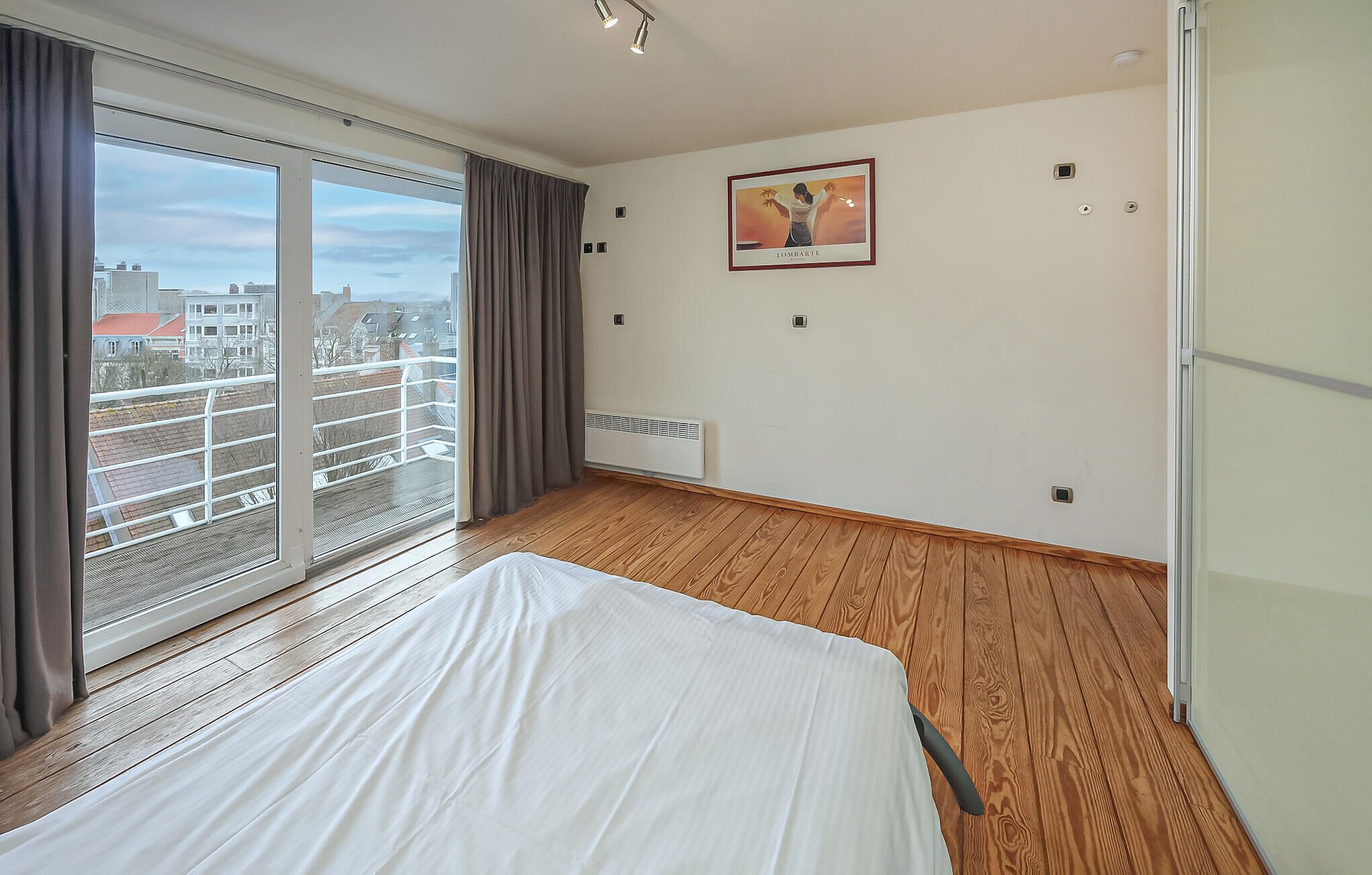 A hotel room with a large window, a bed, a radiator, and a framed picture on the wall.
