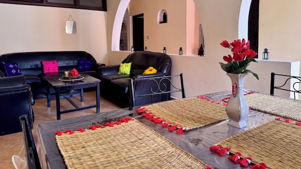 A living room with a black leather sofa, a coffee table, and a dining table with woven placemats and red tassels. - Villa Riad Style (with garden access) - Wifi and Parking (Agadir)