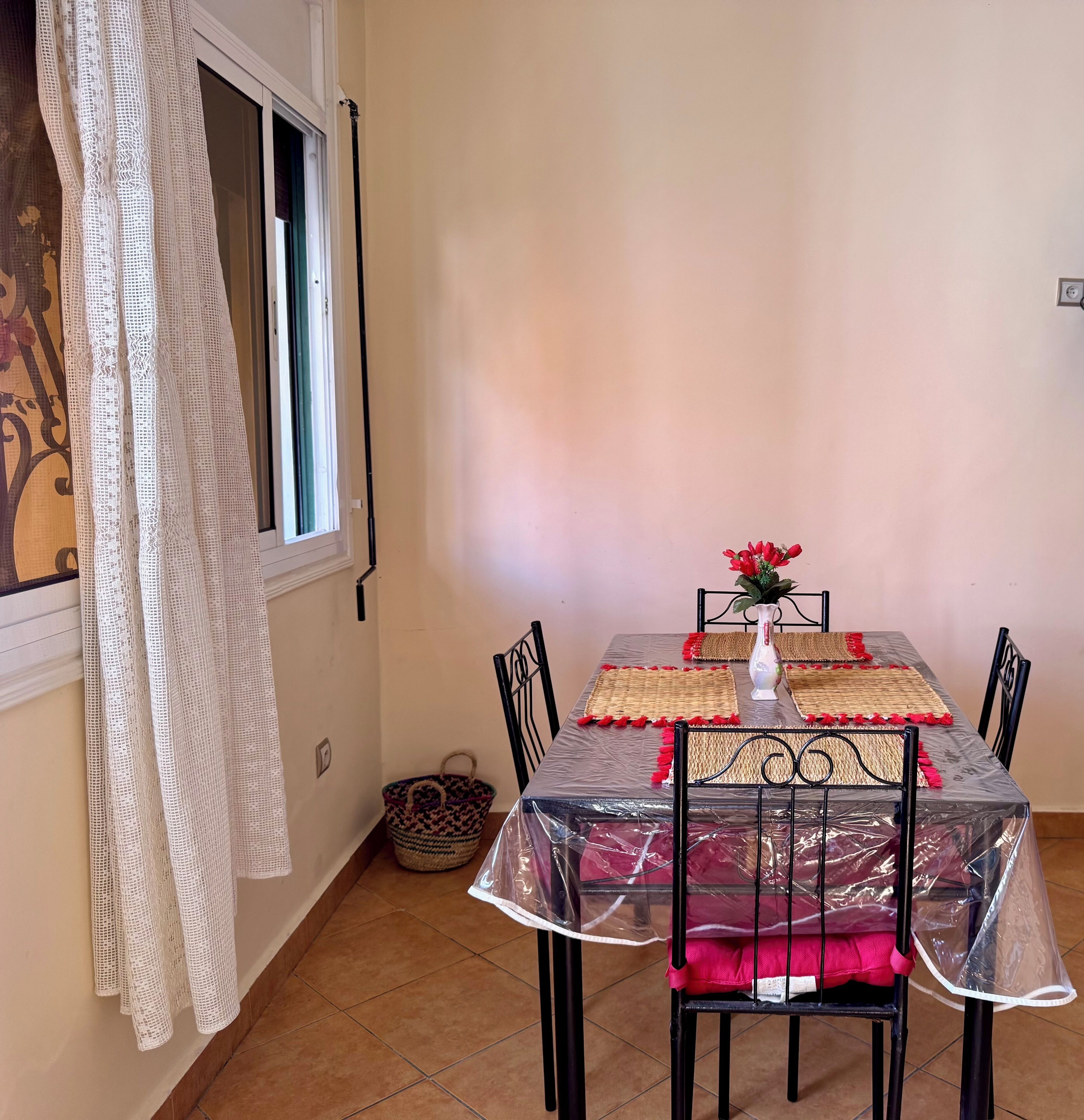 A dining area with a table set for four, covered with a clear plastic tablecloth and surrounded by four chairs. A woven placemat with red tassels is placed under each chair. A vase with red flowers is on the table.