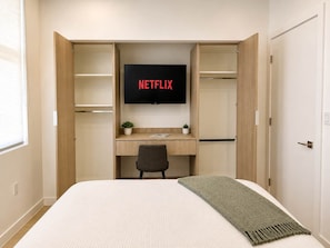 A bedroom with a bed, a desk with a chair, a television mounted above the desk, and a wardrobe with open doors. - South Beach 2BR Apartment: Pool & Bay View #103 (Miami Beach)