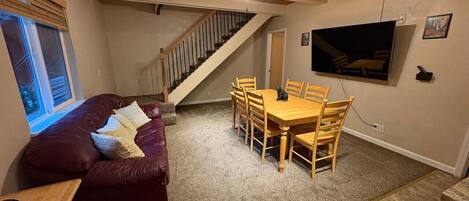 Un salon chaleureux avec un canapé en cuir rouge, une table à manger en bois et des chaises. Un escalier mène à l’étage supérieur.