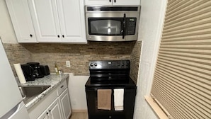 A compact kitchen with white cabinets, a stainless steel microwave, a black stove, and a small countertop with a coffee maker and bottles.