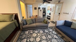 A compact living space with a sofa, two beds, a patterned rug, a ceiling fan, and a kitchen area in the background.