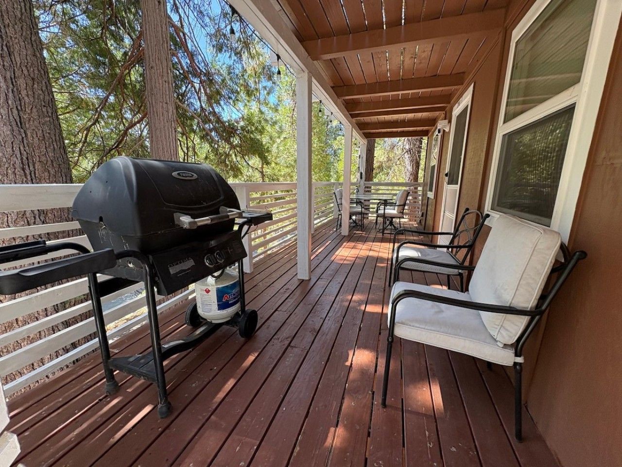 A covered outdoor deck with a grill, chairs, and a view of trees.