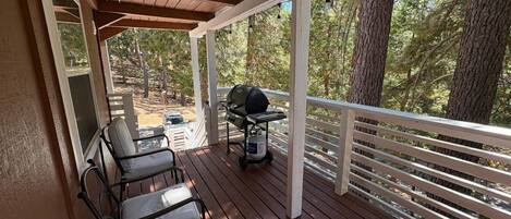 A covered outdoor deck with wooden flooring, metal chairs, a grill, and a view of a forested area.
