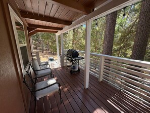 A covered outdoor deck with wooden flooring, metal chairs, a grill, and a view of a forested area.