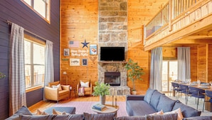 Interior acogedor de una cabaña con chimenea de piedra, un sofá grande, zona de comedor y balcón.