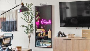 Ein modernes Wohnzimmer mit einem großen Flachbildfernseher, einem Spiegel mit einer Neonreklame, einer Pflanze und einer Holzkassette mit einer Willkommensanzeige.