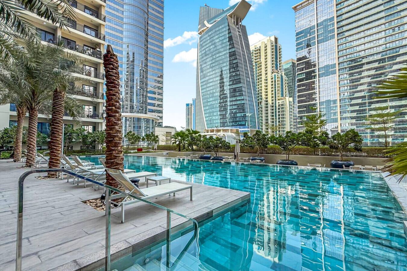 A swimming pool with lounge chairs and palm trees, surrounded by high-rise buildings.