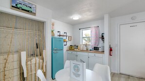 A compact kitchen with a teal refrigerator, white chairs, a small dining table, and a window with a curtain. - Pineapple Shores Retro Inn 2 (Daytona Beach)