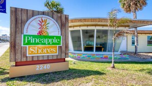 A sign for Pineapple Shores with a beach-themed logo, a modern building with large windows, and a colorful mosaic pathway. - Pineapple Shores Retro Inn 2 (Daytona Beach)
