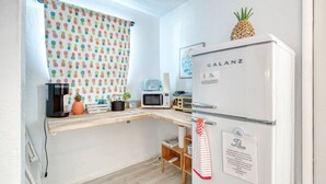 Eine kleine Küche mit einem weißen Kühlschrank, einer Mikrowelle, einer Kaffeemaschine und einem Regal mit einer Ananas und einer Pflanze.