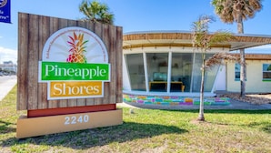 A sign for Pineapple Shores with a beach-themed logo, a modern building with large windows, and a colorful mosaic pathway. - Pineapple Shores Retro Inn 3 (Daytona Beach)