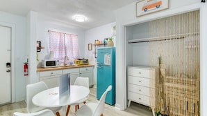 A compact kitchen with a microwave, toaster, and a blue refrigerator. There is a dining table with white chairs and a small window with patterned curtains. - Pineapple Shores Retro Inn 3 (Daytona Beach)
