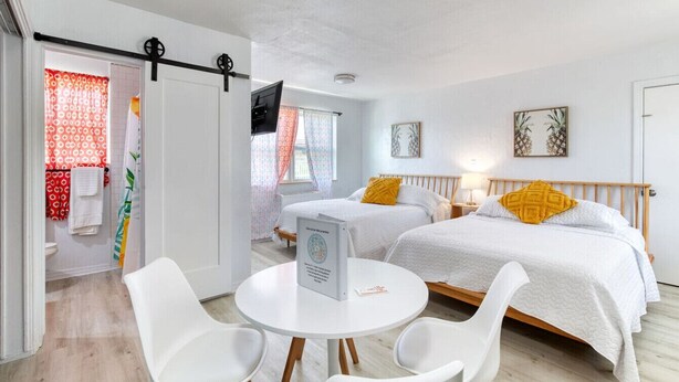 A hotel room with two beds, a round table with chairs, a TV, and a bathroom with a shower curtain. - Pineapple Shores Retro Inn 5 (Daytona Beach)