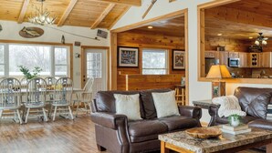 A spacious living room with a brown leather sofa, dining area with white chairs, wooden ceiling beams, and a kitchen area with wooden cabinets. - Mins to Hunter Mountain Sleeps 36 Pool, Spa, Games & Bar Cozy Peaks Lodge by Avantstay (Tannersville)