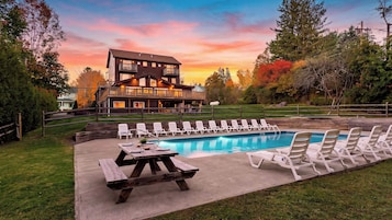 Un área junto a la piscina con sillones reclinables y una mesa de picnic, rodeada por una terraza de madera y un edificio grande con varios balcones.