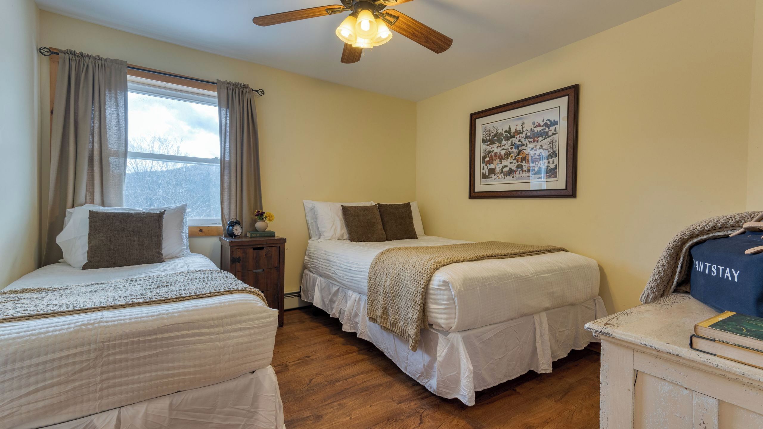A room with two beds, a ceiling fan, a framed picture on the wall, and a nightstand with a clock and flowers.
