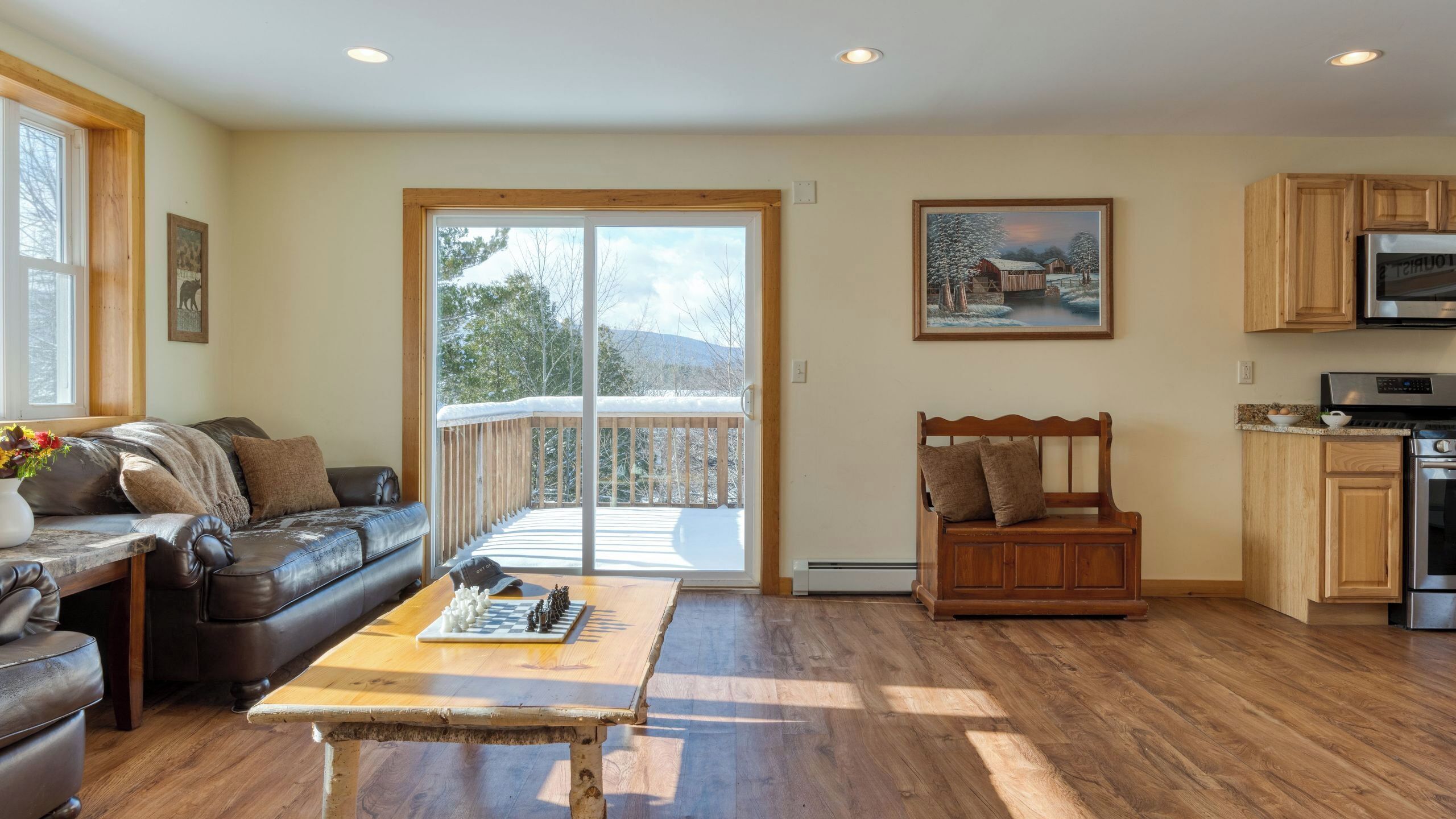 A cozy living room with a fireplace, a wooden coffee table, and a sofa. There is a sliding glass door leading to a balcony with a view of trees and mountains.