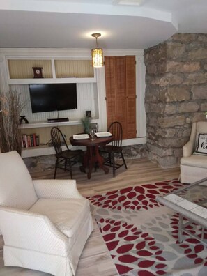 A living room with a stone wall, a dining table with chairs, a television, and a fireplace. - The Garden Suite (Eureka Springs)