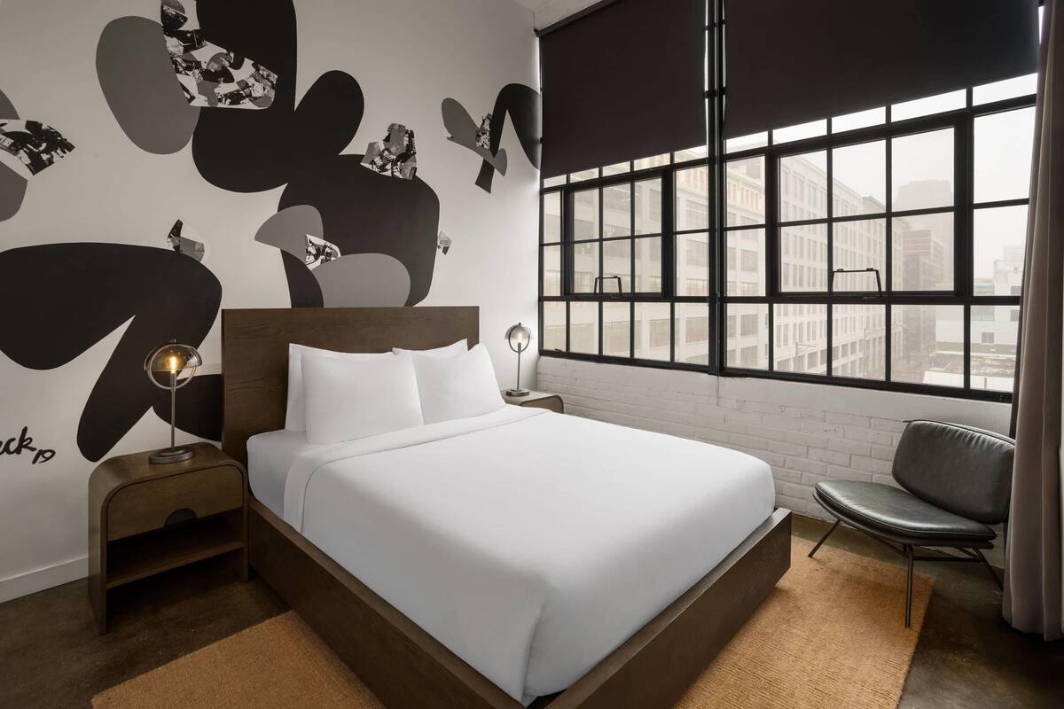 A modern hotel room with a large bed, a nightstand with a lamp, a chair, and a large window with a city view.