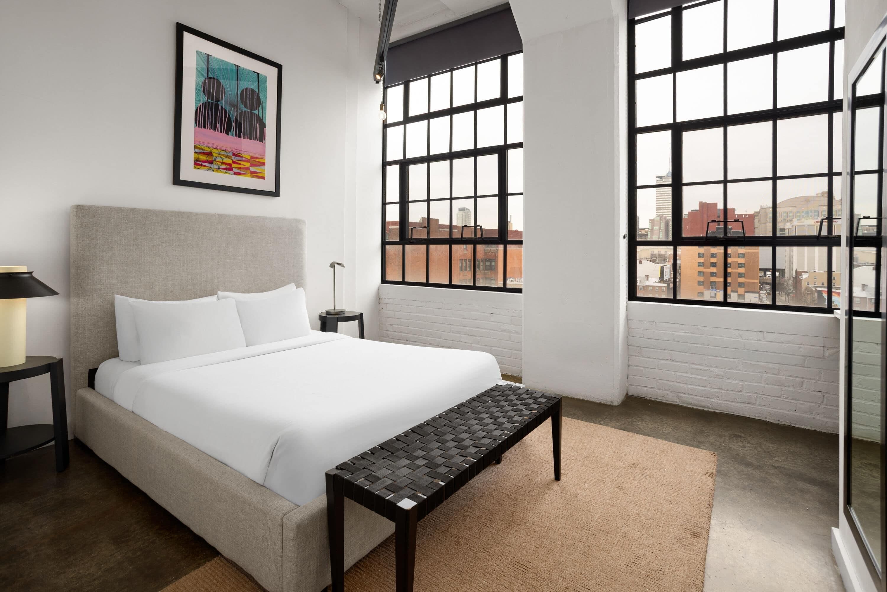 Un dormitorio moderno con cama, banca, mesita de noche, lámpara y vistas a la ciudad a través de amplias ventanas.