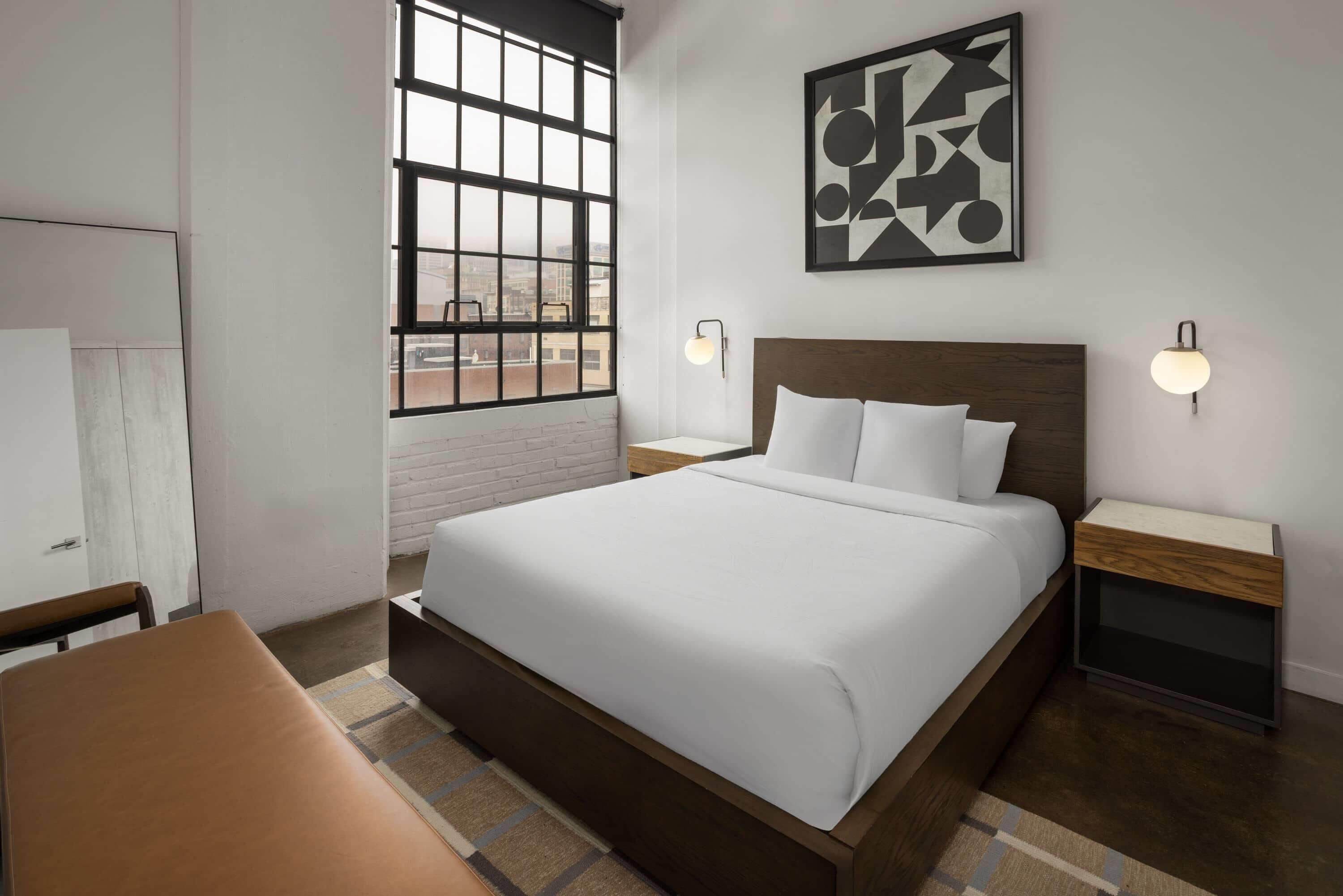 Ein modernes Hotelzimmer mit einem großen Bett, zwei Nachttischen, einem Fenster mit Blick und einem geometrischen Wandbild.
