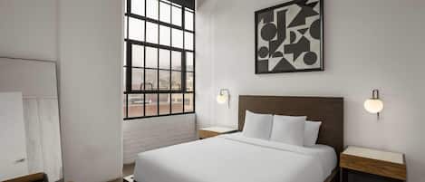 A modern hotel room with a large bed, two bedside tables, a window with a view, and a geometric wall art piece.