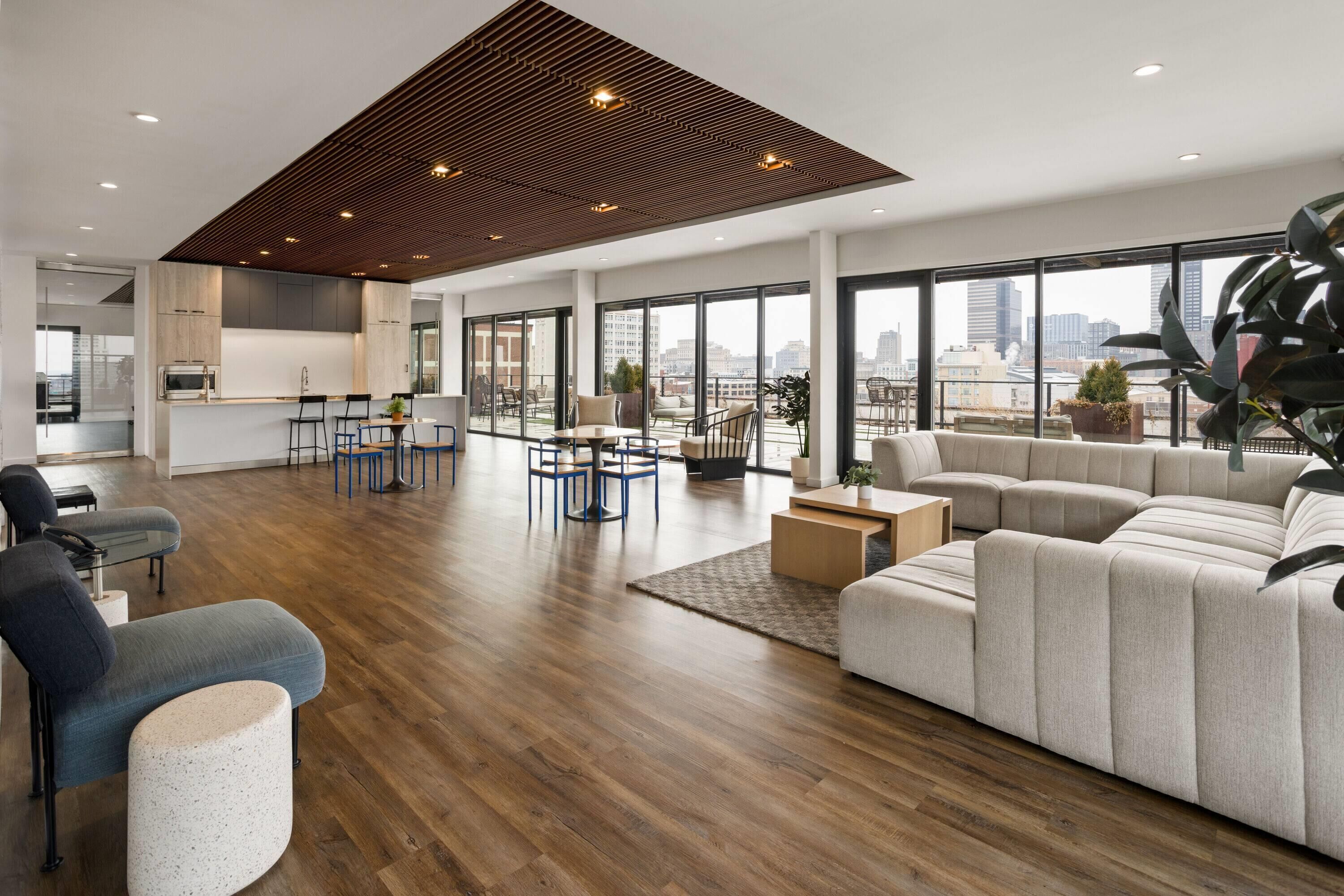Uma sala espaçosa com um sofá grande, uma mesa de jantar e cadeiras, uma cozinha ao fundo e vista para a cidade através de amplas janelas.