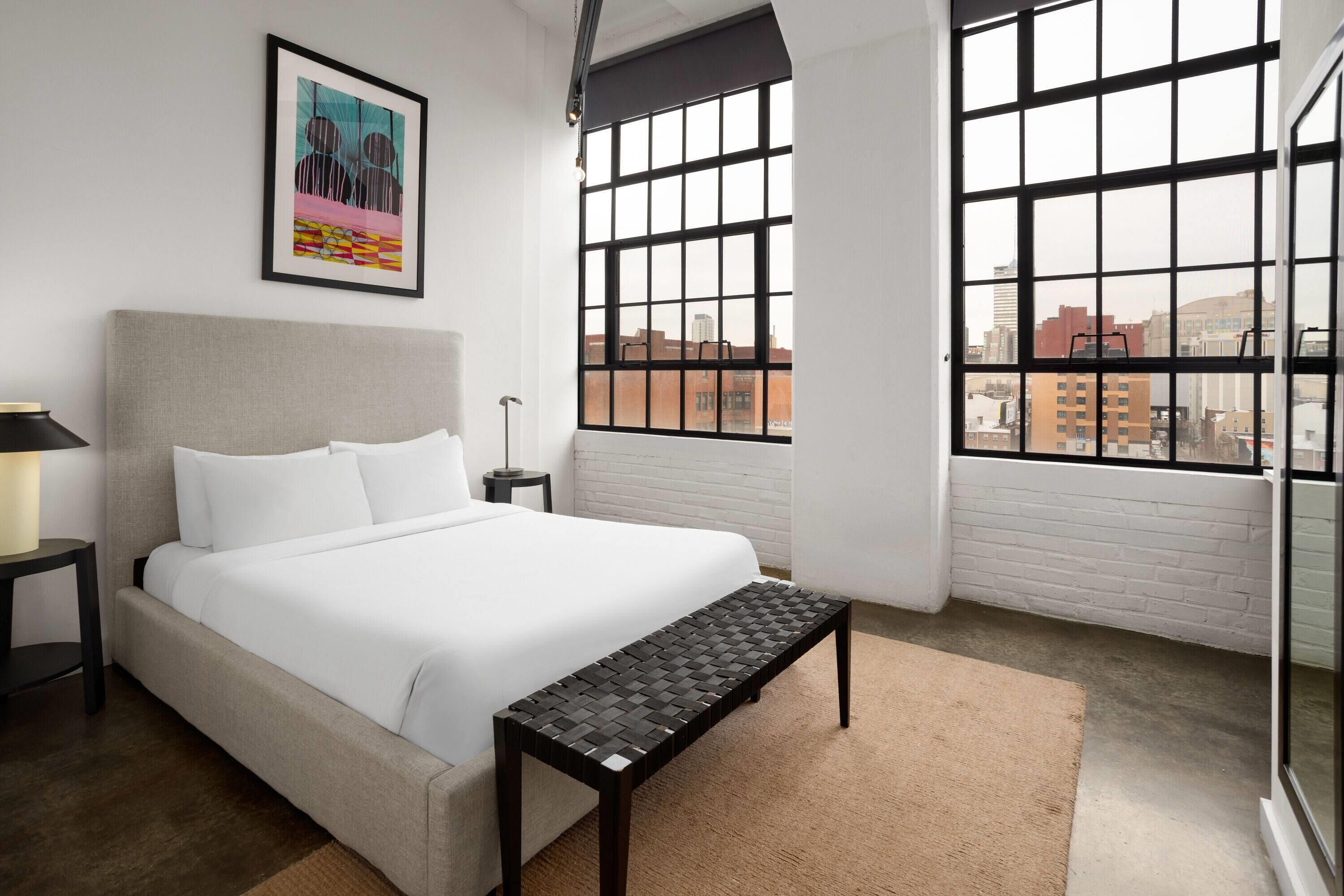Un dormitorio con cama, un banco, una mesita de noche, una lámpara y una ventana con vistas a la ciudad.