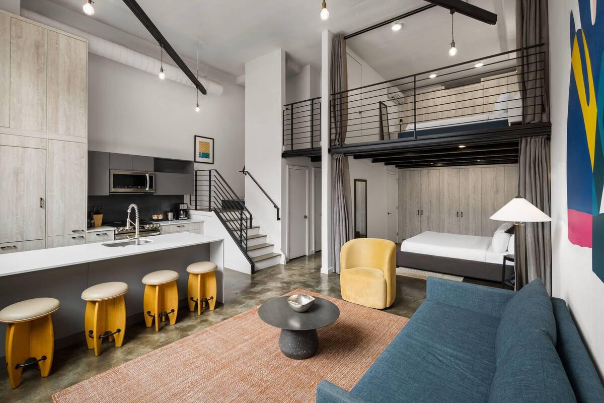 A modern interior with a kitchen, dining area, and living space, featuring a staircase, a sofa, and a yellow armchair.