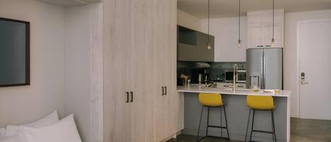 A modern hotel room with a bed, a small kitchenette, and two yellow bar stools.
