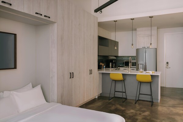 Una habitación de hotel moderna con una cama, un pequeño rincón de cocina y dos taburetes amarillos.