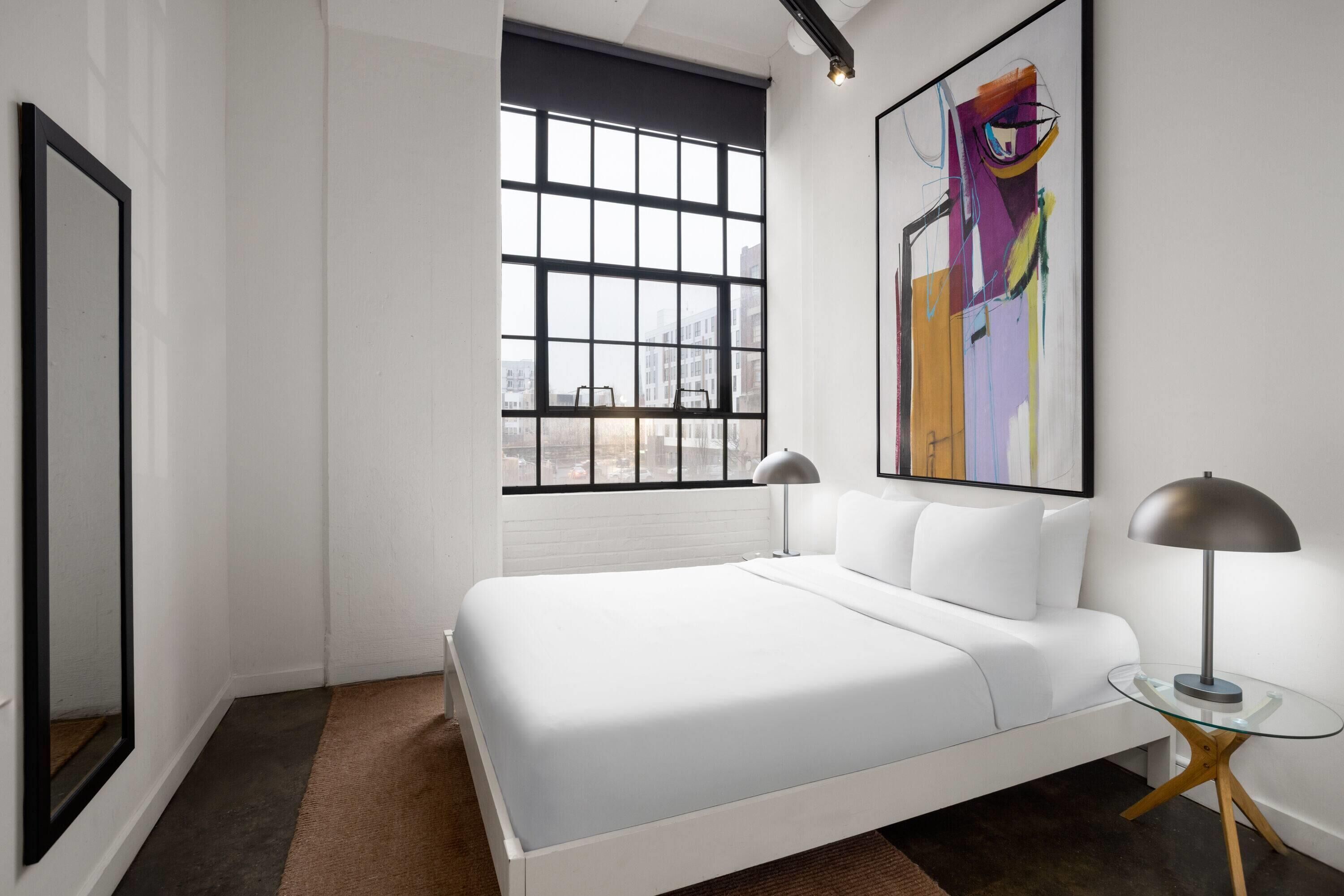 Un dormitorio moderno con un ventanal grande, una cama, una mesita de vidrio y una lámpara.